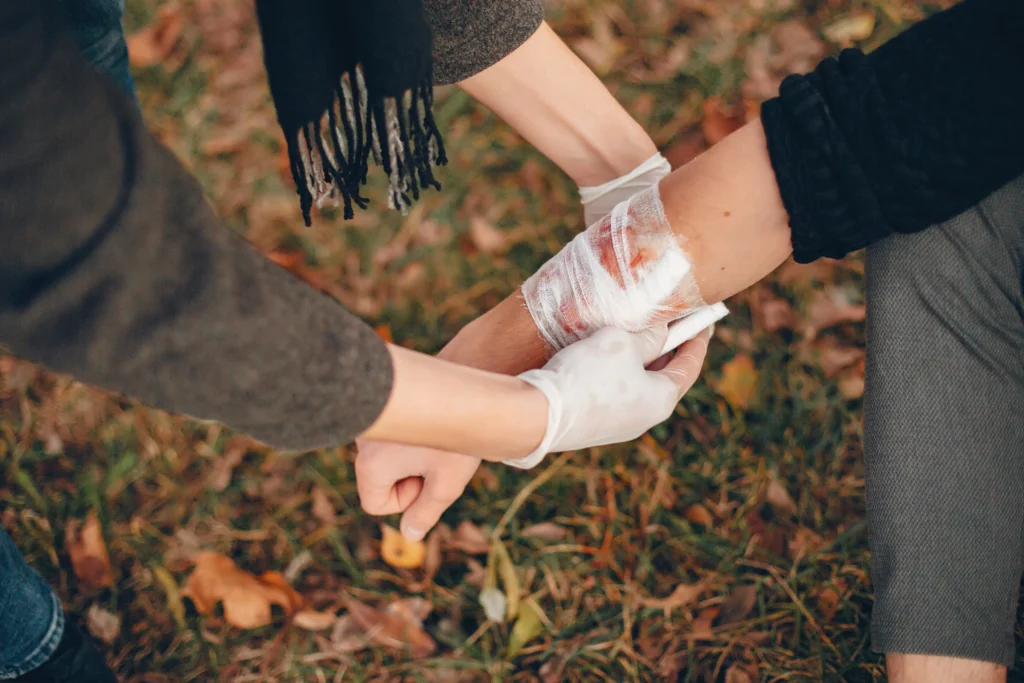 A person in gloves cares for a bandaged arm with an injury, while another person holds their hand for support on a grassy surface.