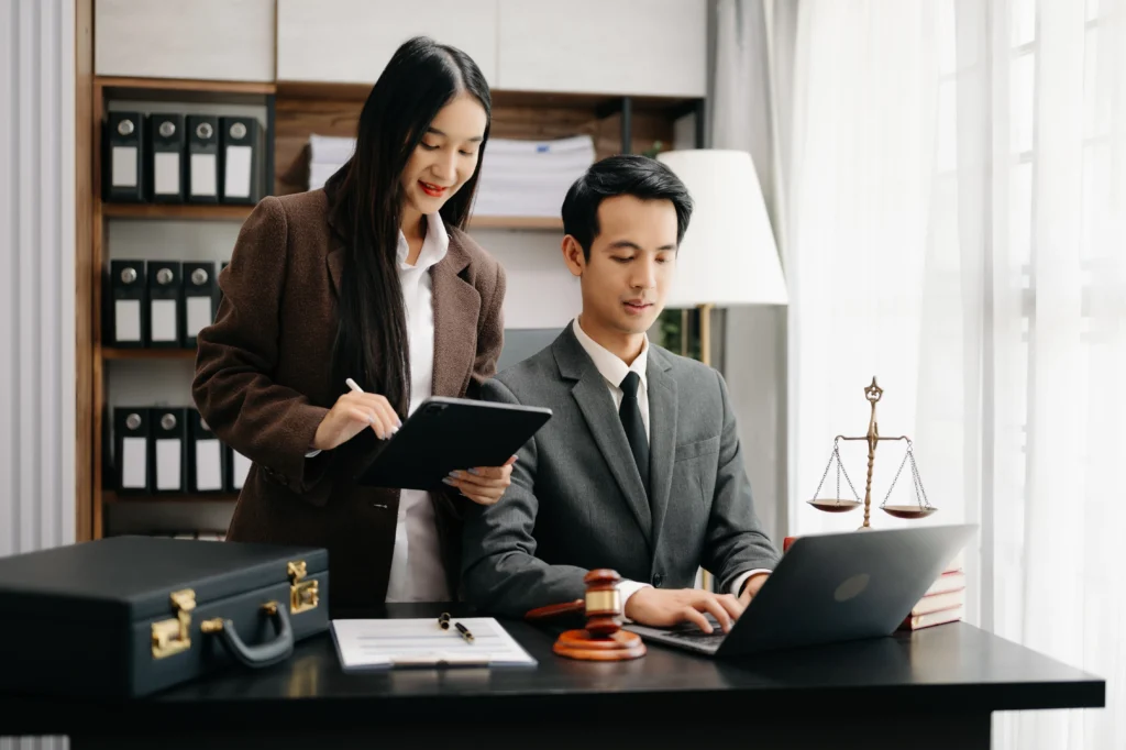 A woman in a brown blazer stands by a man in a suit, reviewing notes on a tablet at a stylish office desk with legal symbols.