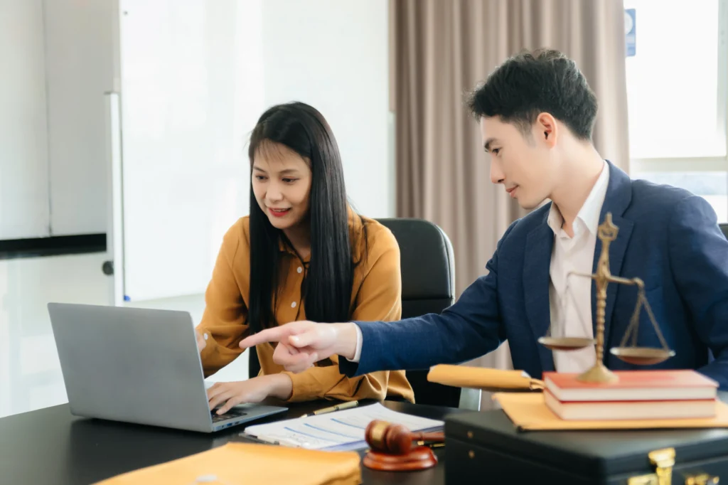 A man in a suit gestures while discussing with a woman in an orange blouse, both focused on a laptop amidst legal documents and a gavel.