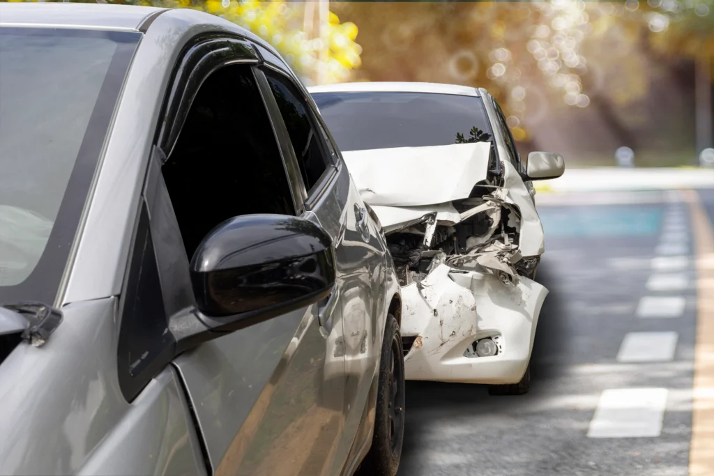 A silver car parked next to a white car with a damaged rear, indicating a recent collision, set on a blurred tree-lined road.