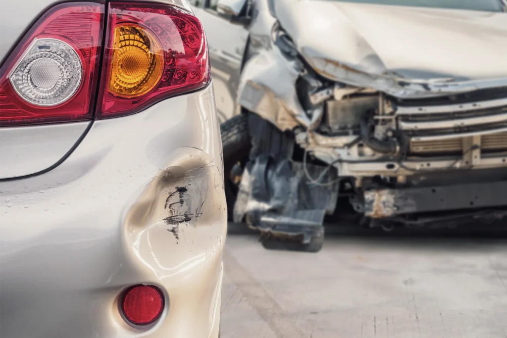 A close-up of a dented silver car bumper with a damaged red taillight, while a severely wrecked vehicle is blurred in the background.
