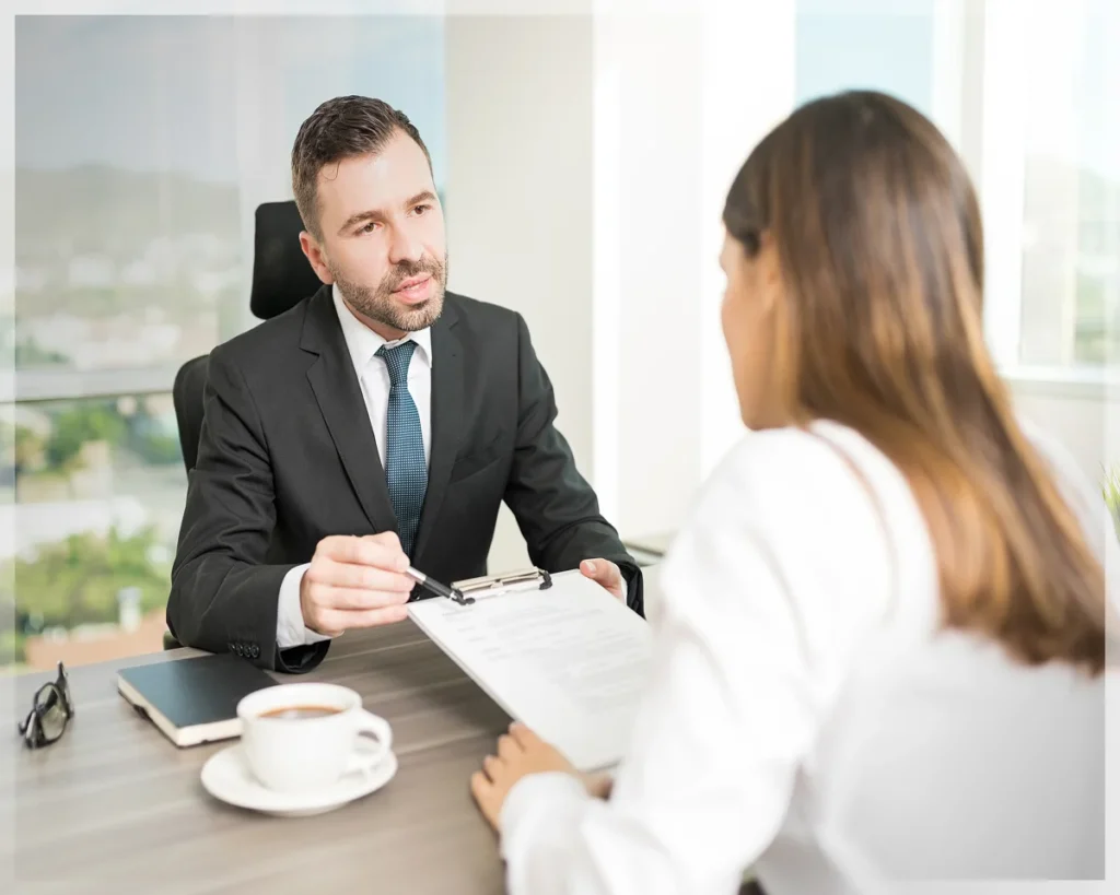 A professional interview setting with a man in a suit handing a clipboard to a woman seated across from him, coffee cup on the table.