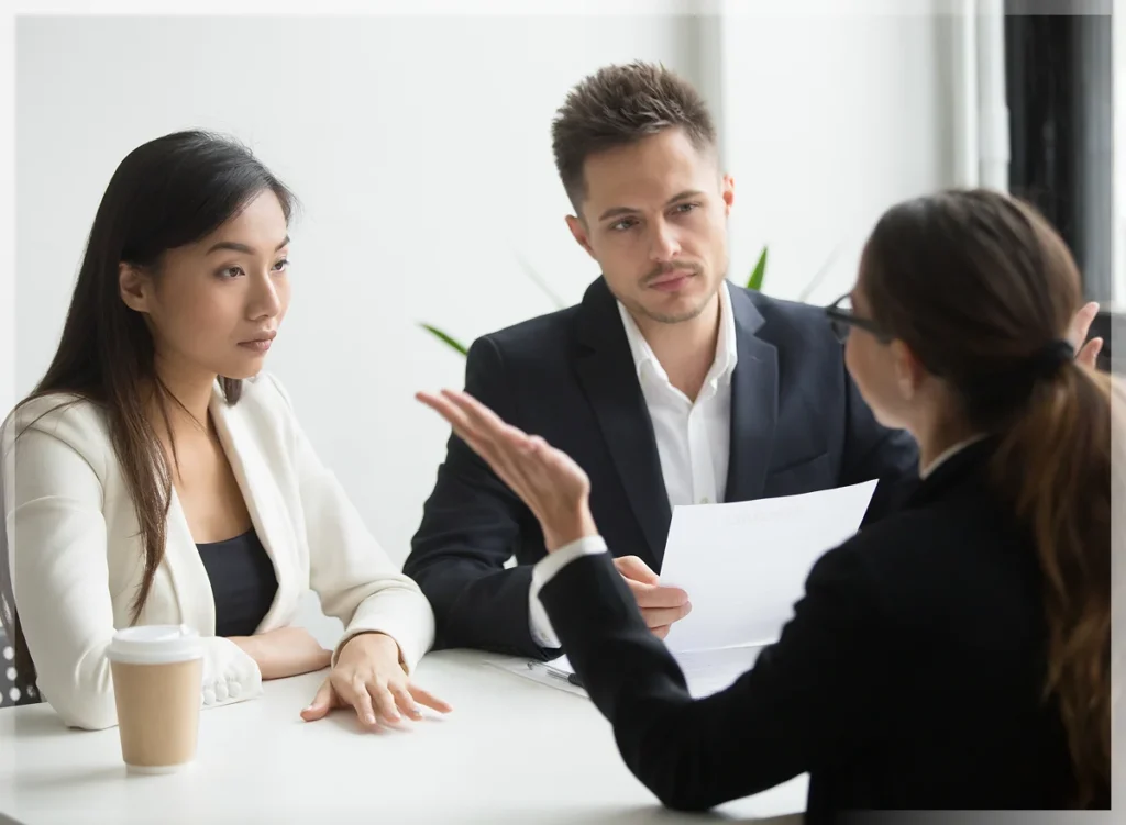 A professional meeting in a bright office setting, with two individuals attentively listening to a speaker holding documents.