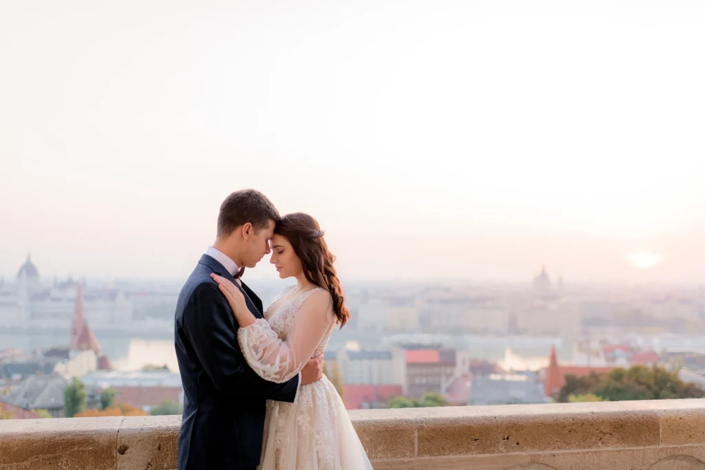 A man and woman in wedding attire embracing, with the sky in the background. The woman is wearing a wedding dress and veil, while the man is dressed in a suit. Their expressions convey love and romance during an outdoor ceremony.