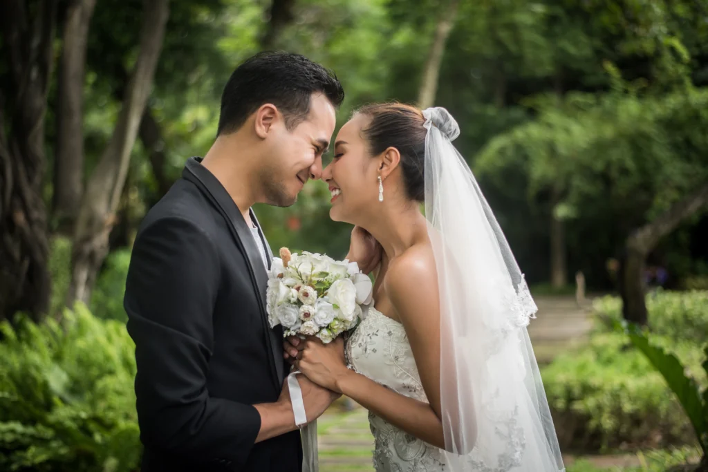 A man and woman in wedding attire share a kiss outdoors, surrounded by trees. The woman is wearing a bridal gown and veil, while the man is dressed in a suit. The scene captures a romantic moment during their wedding ceremony.